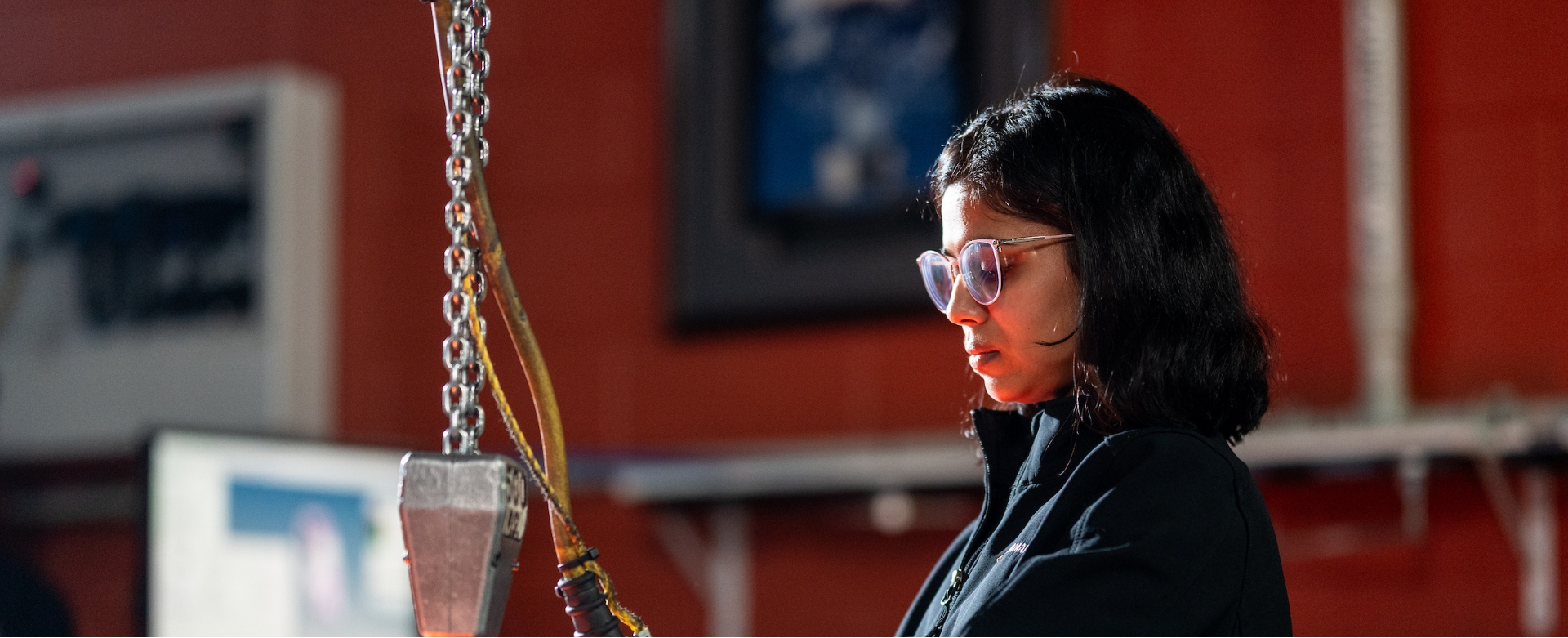 Woman with glasses focused on her work in an indoor industrial setting with machinery in the background.