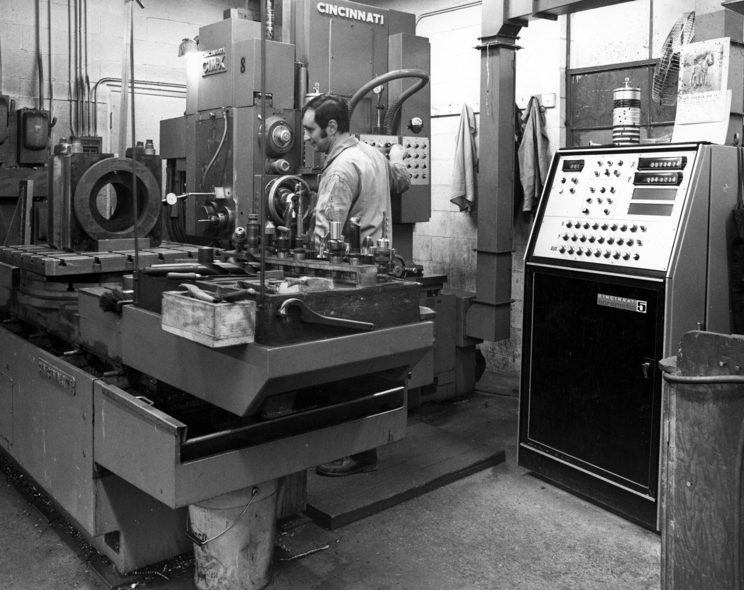 Black and white photo of a man operating industrial machinery in a factory workshop with control panels and equipment surrounding him.