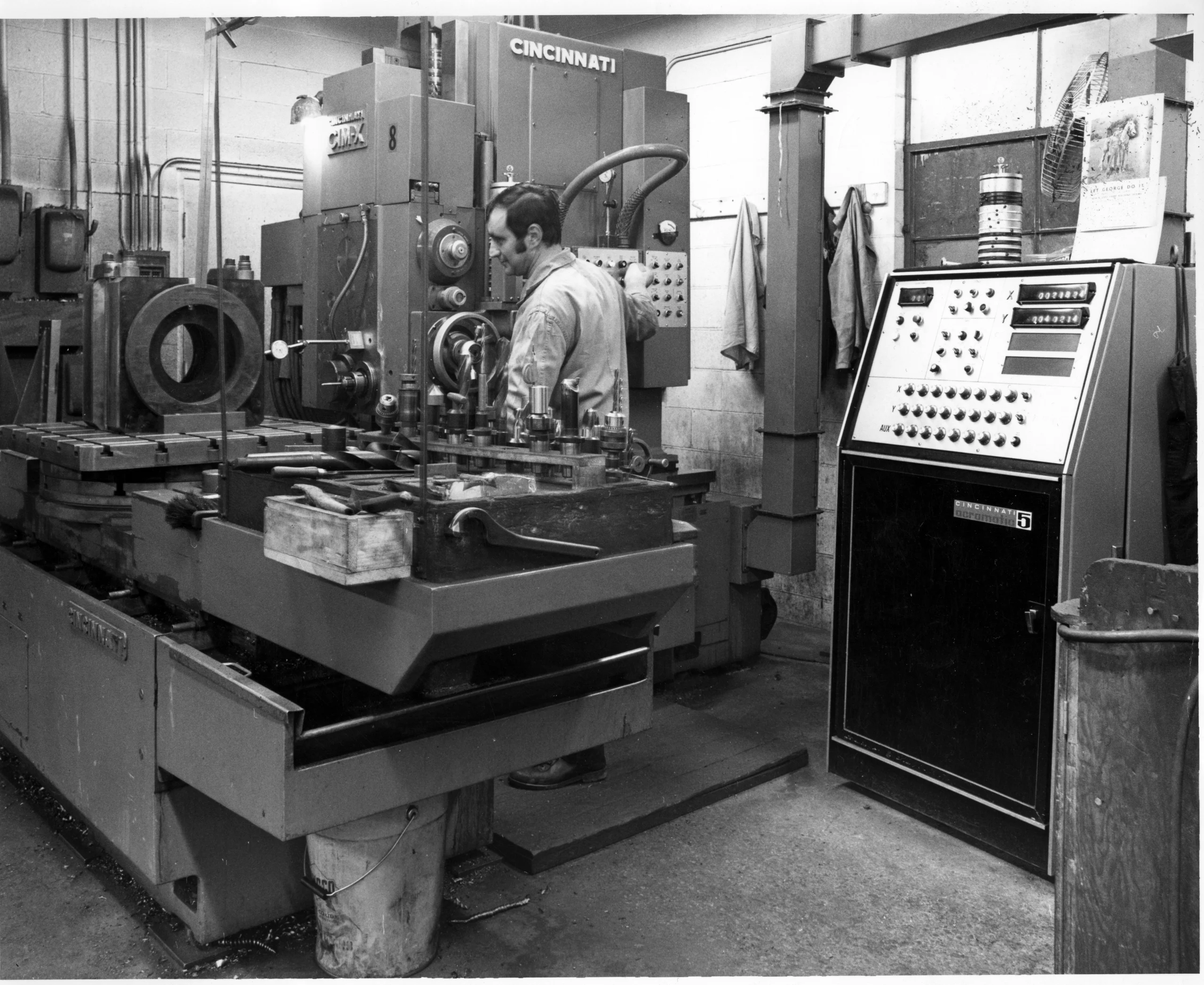 Black and white photo of a man operating industrial machinery in a factory workshop with control panels and equipment surrounding him.