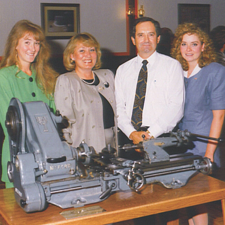 Four professionals standing behind a metal lathe machine in an industrial workshop setting.