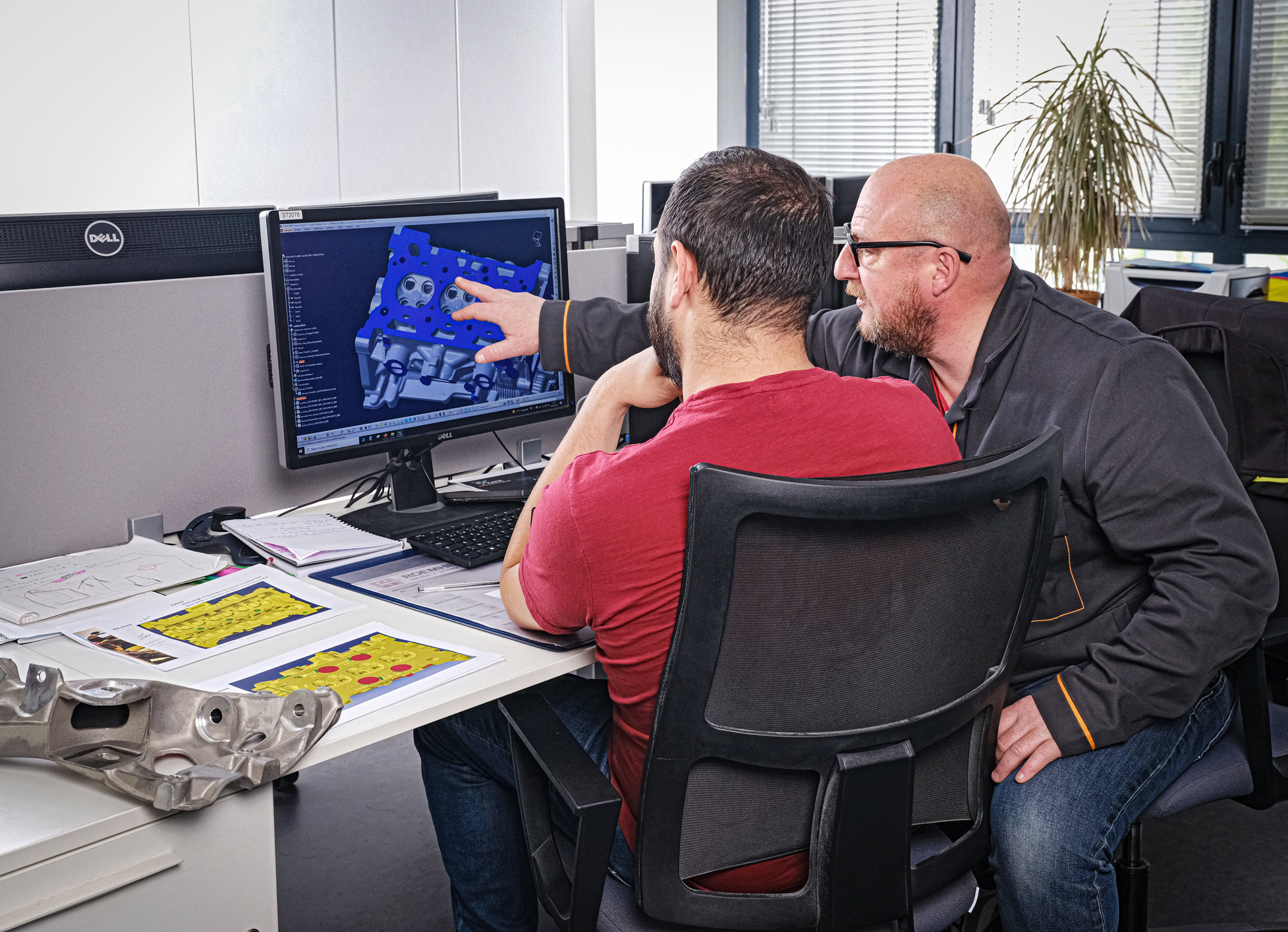 Two men collaborate and analyze 3D models on a computer screen in a modern office workspace.