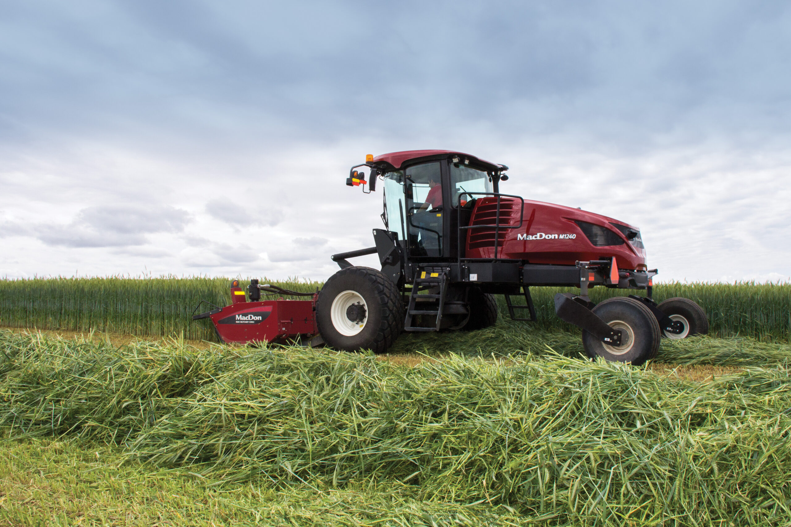 Red agricultural mower cutting grass in a large green field under a cloudy sky.