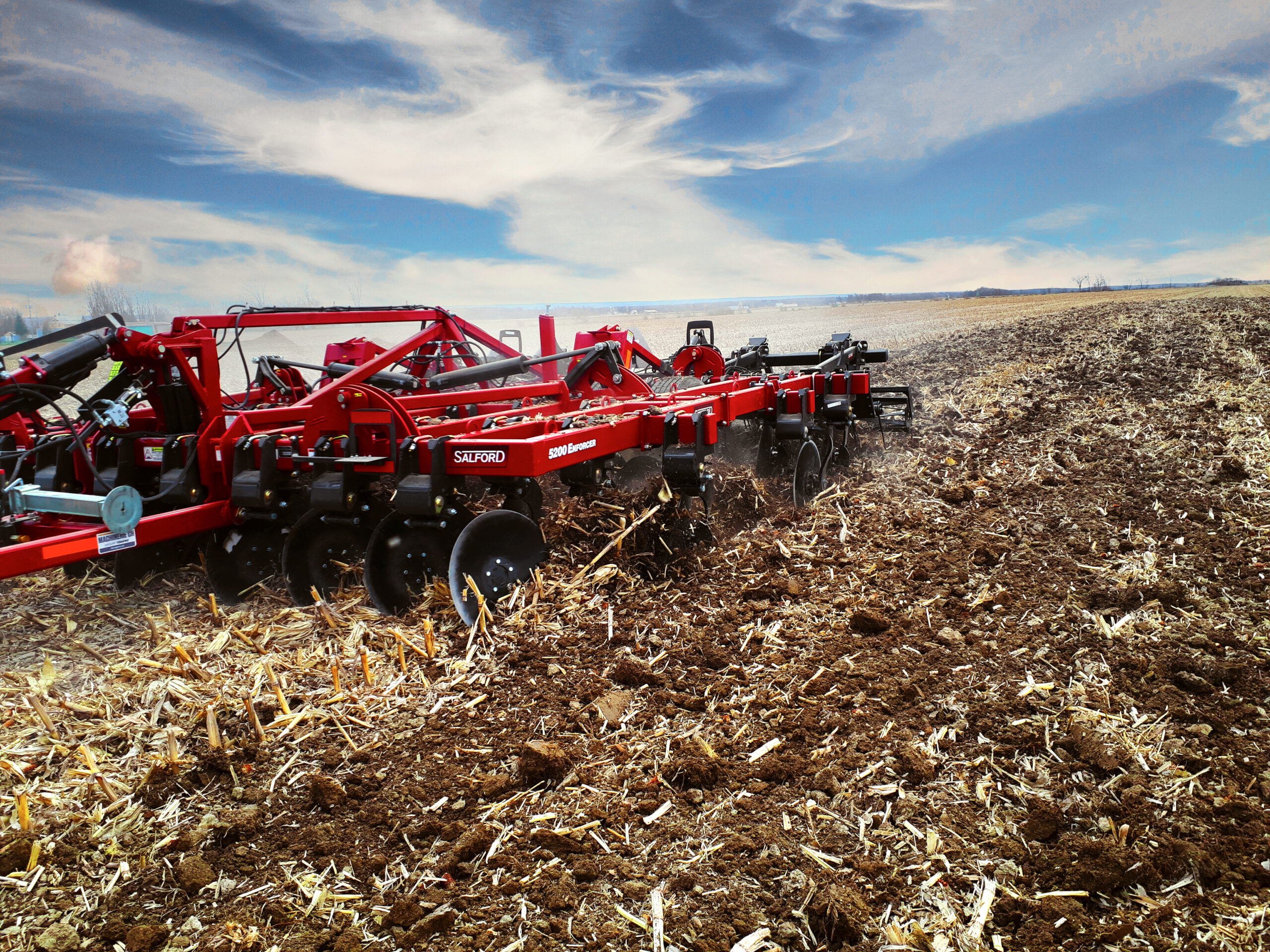 Red agricultural plow tilling dry soil in a vast field under a partly cloudy sky.
