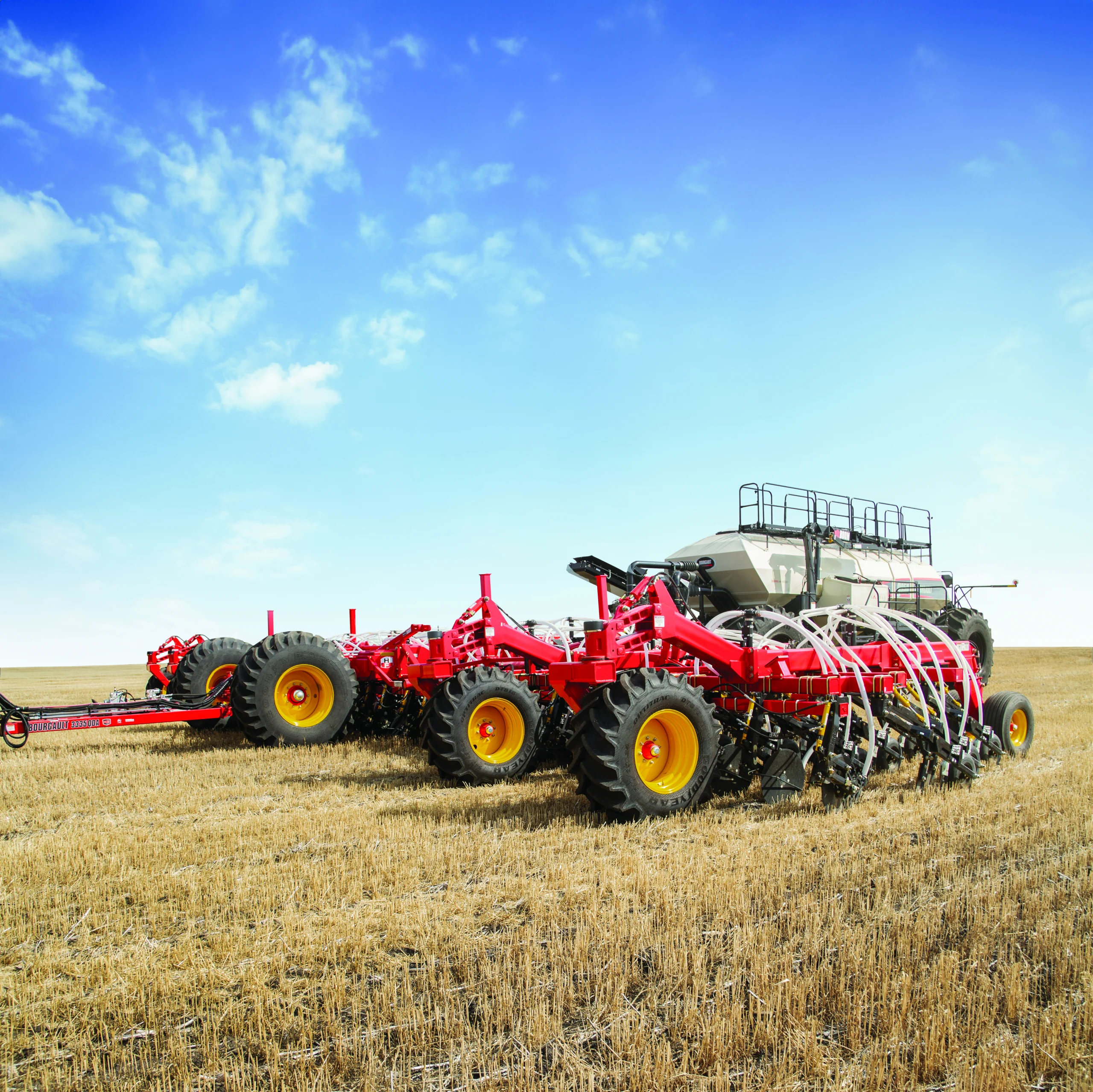 Red agricultural planter machine with multiple seed drills working on a harvested field under a clear blue sky.