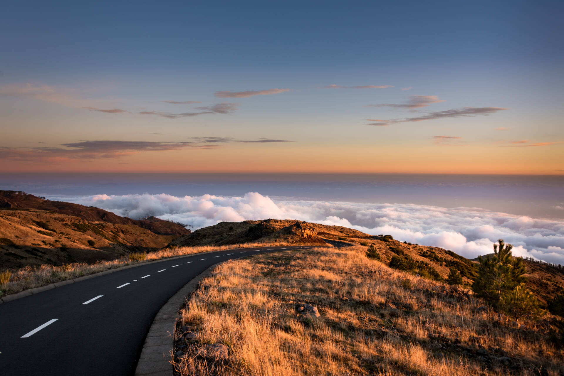 Scenic mountain road curving through golden grasslands above a blanket of clouds during a colorful sunset.