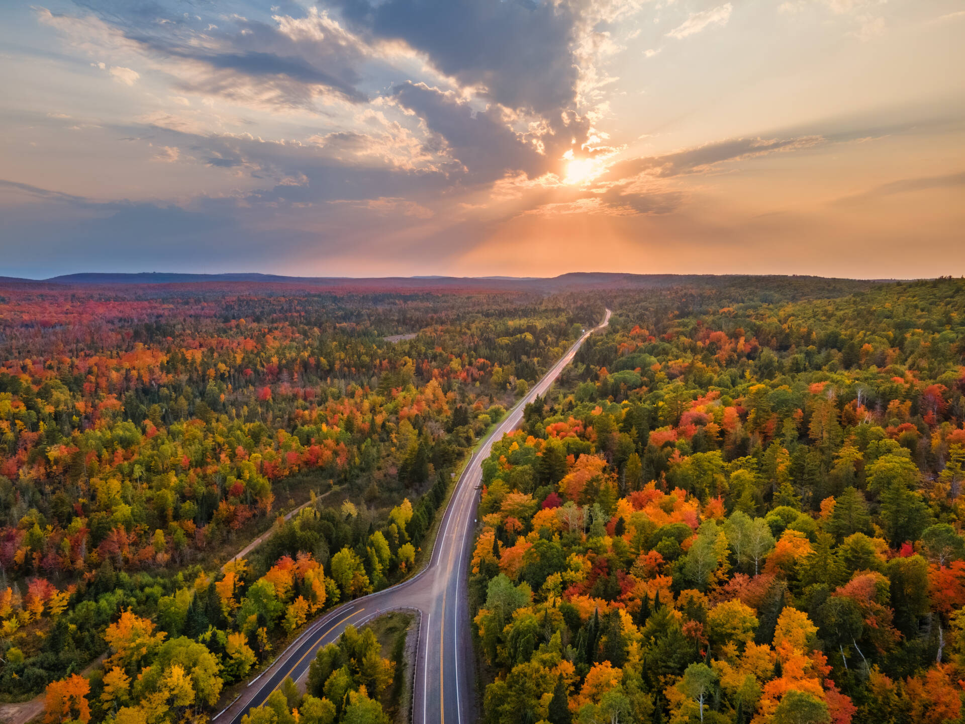 A scenic aerial view of a winding road cutting through a dense forest with vibrant autumn foliage under a dramatic sunset sky.