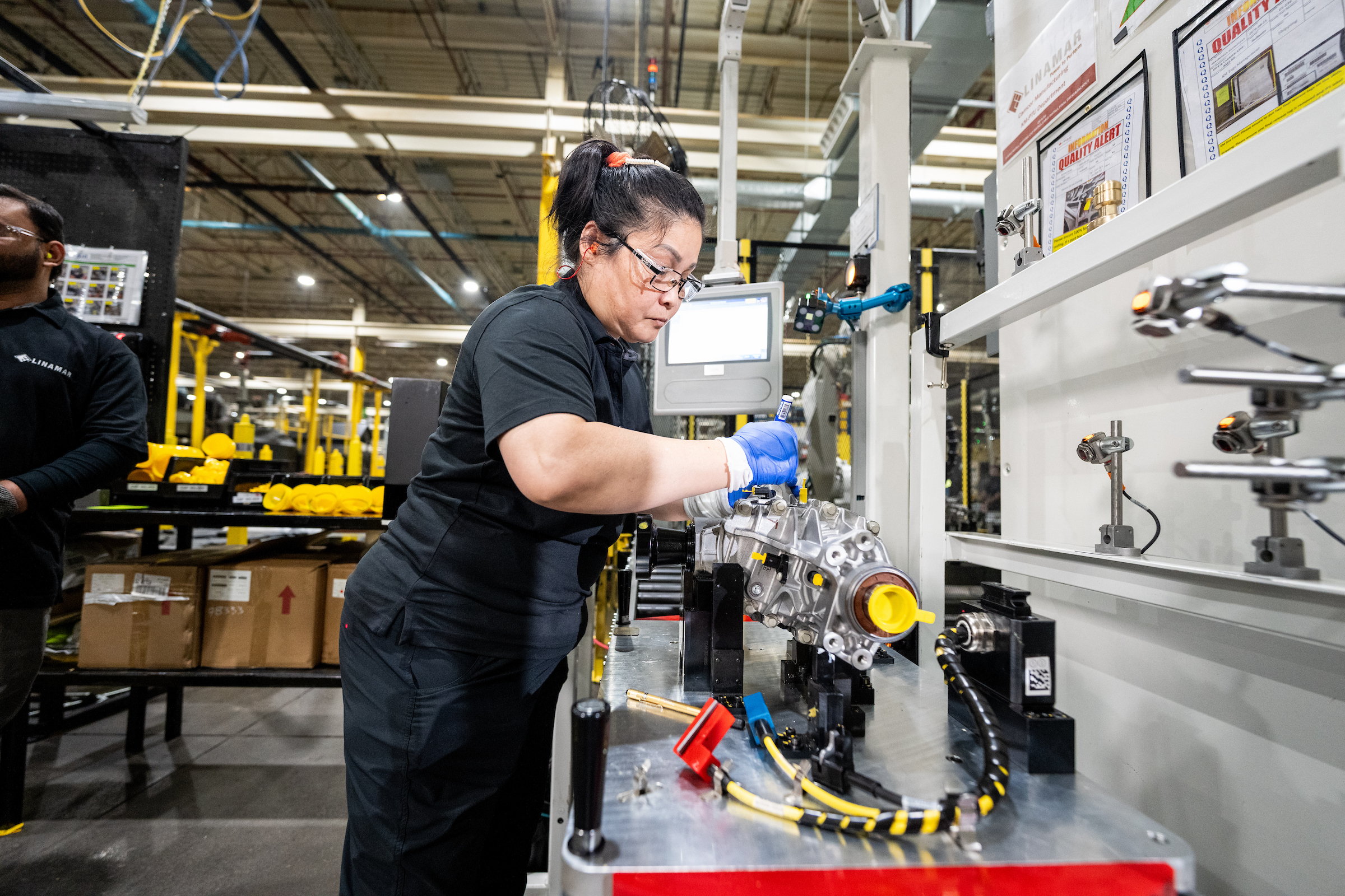 Factory worker wearing gloves and safety glasses operating machinery on an industrial assembly line inside a manufacturing plant.
