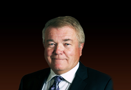 Professional headshot of a middle-aged man wearing a dark suit and patterned tie against a dark gradient background.