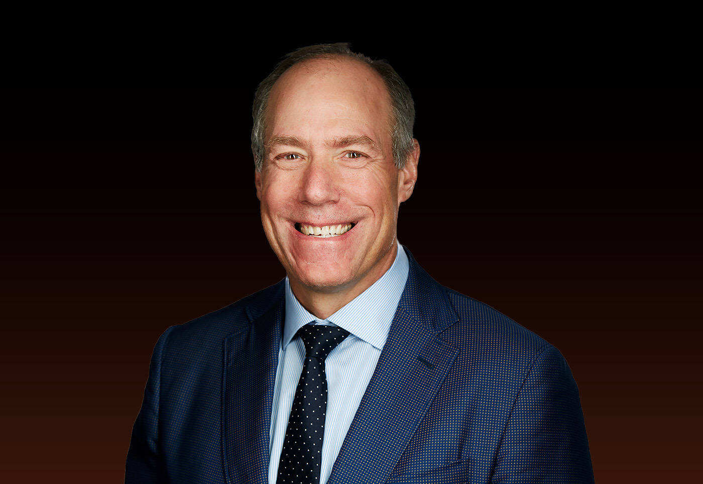 Professional headshot of a smiling middle-aged man wearing a navy blue suit, white shirt, and dark tie against a dark gradient background.