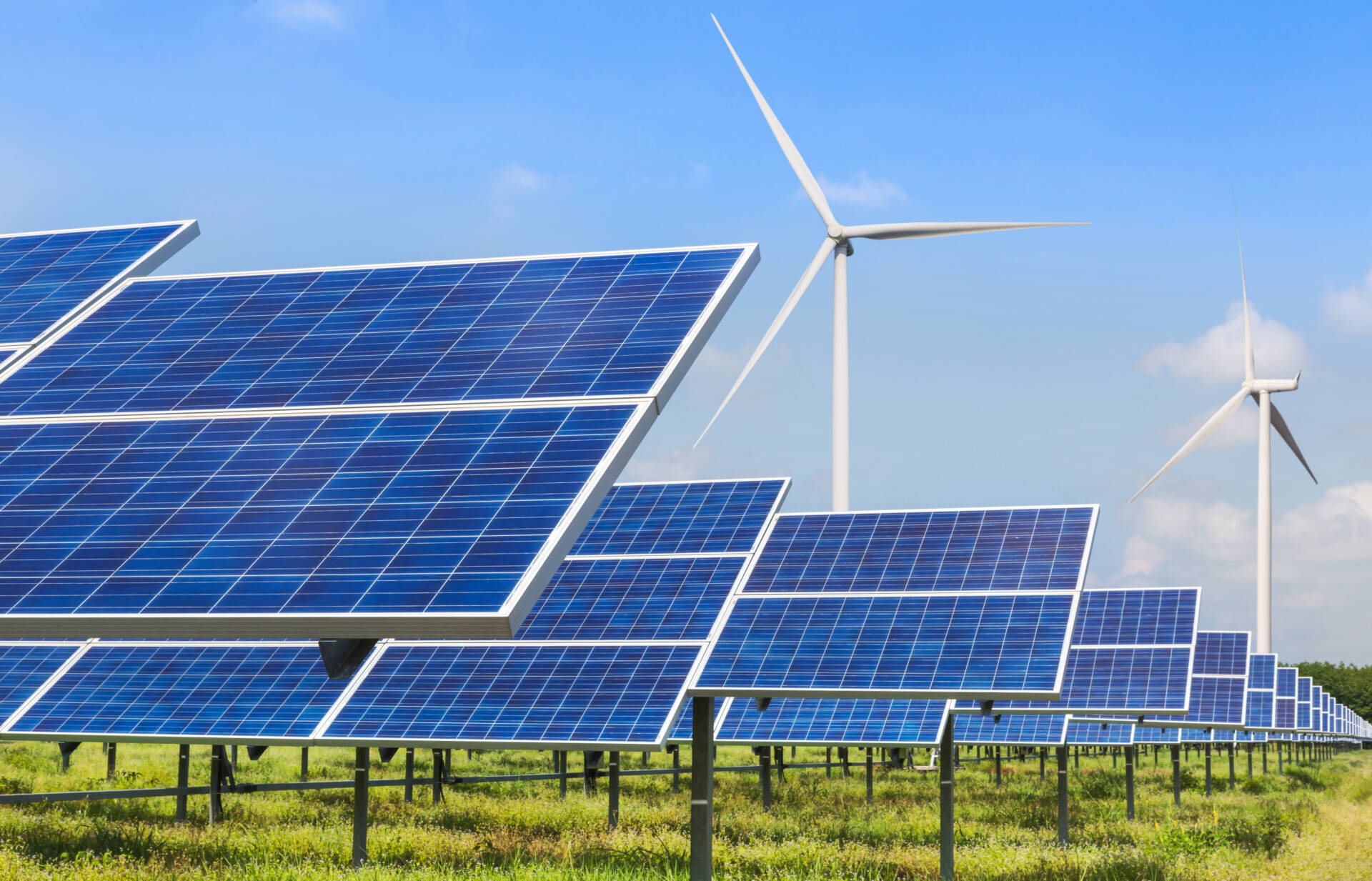 Solar panels installed in a field with wind turbines in the background under a clear blue sky, representing renewable energy sources.
