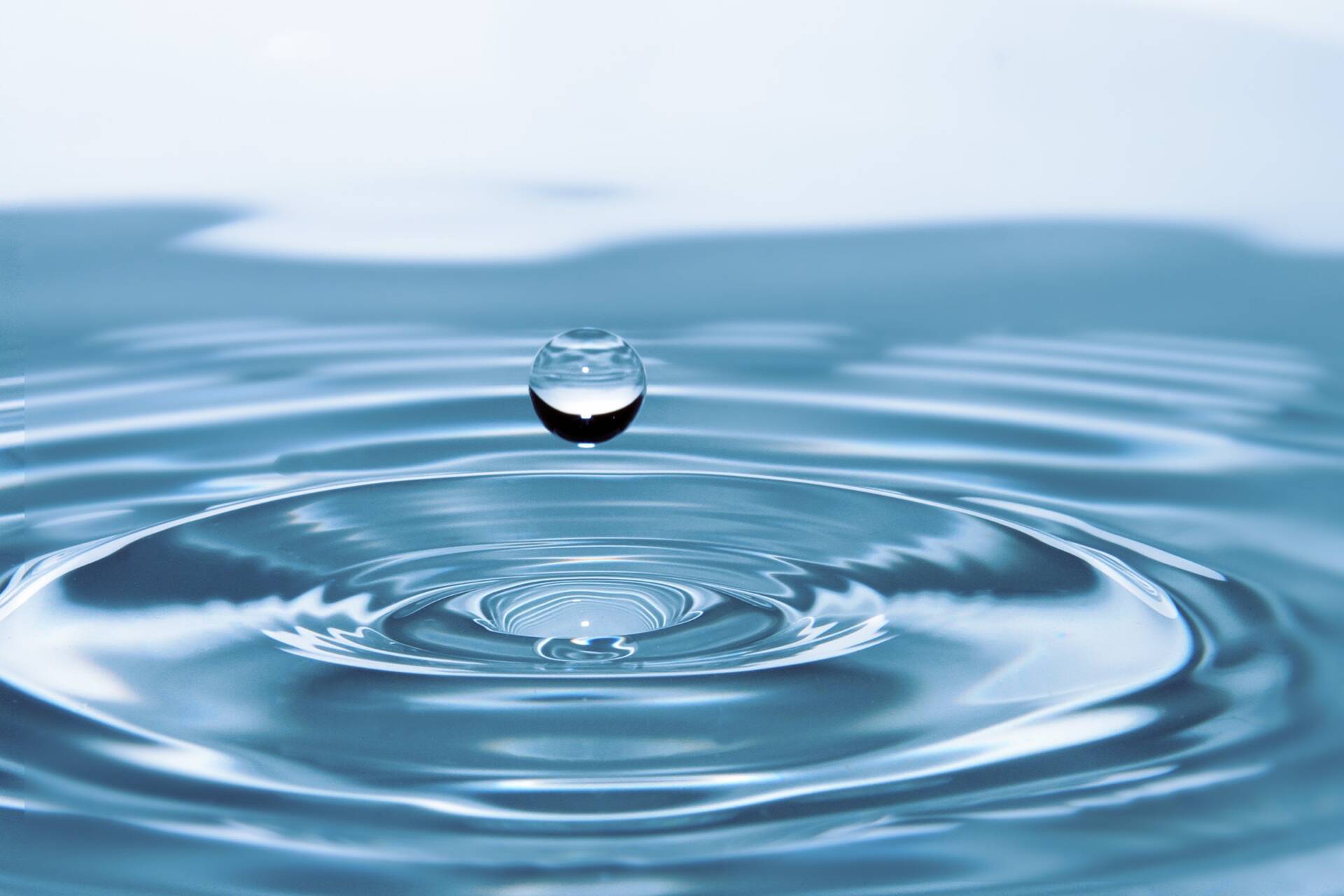Close-up of a water droplet suspended above a calm surface, creating concentric ripples in clear blue water.