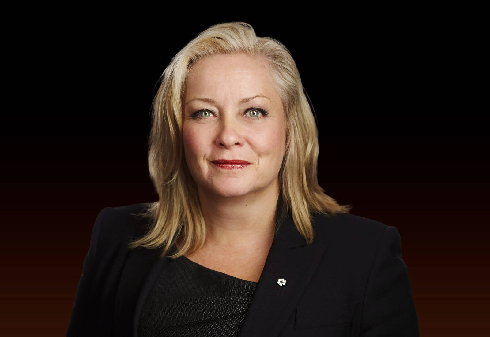 Portrait of a confident woman with blonde hair wearing a black blazer against a dark background.