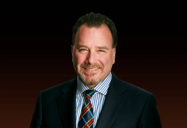 Professional headshot of a smiling man in a dark suit and striped tie against a dark gradient background.