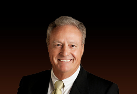 Professional headshot of a smiling middle-aged man wearing a black suit and yellow tie against a dark brown background.