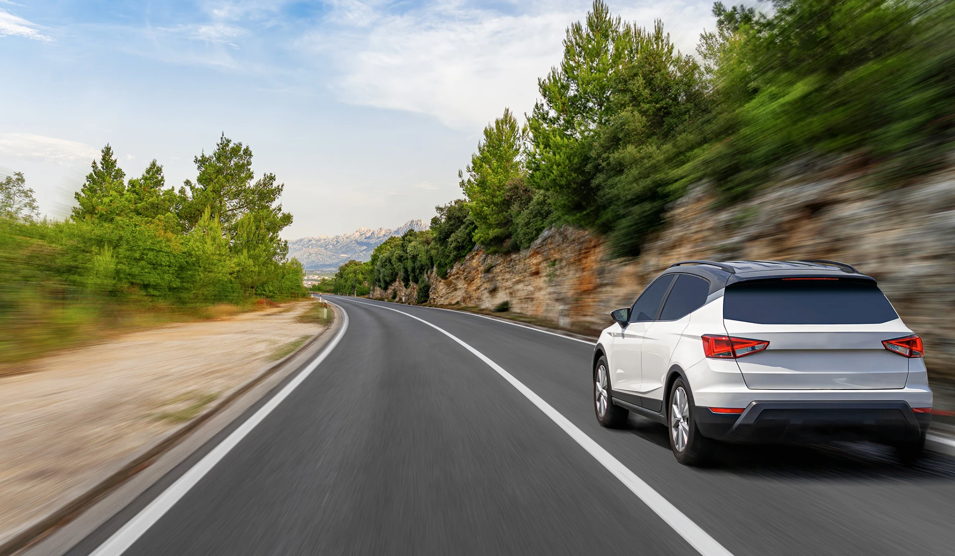 White SUV driving on a winding mountain road surrounded by pine trees under a partly cloudy sky.