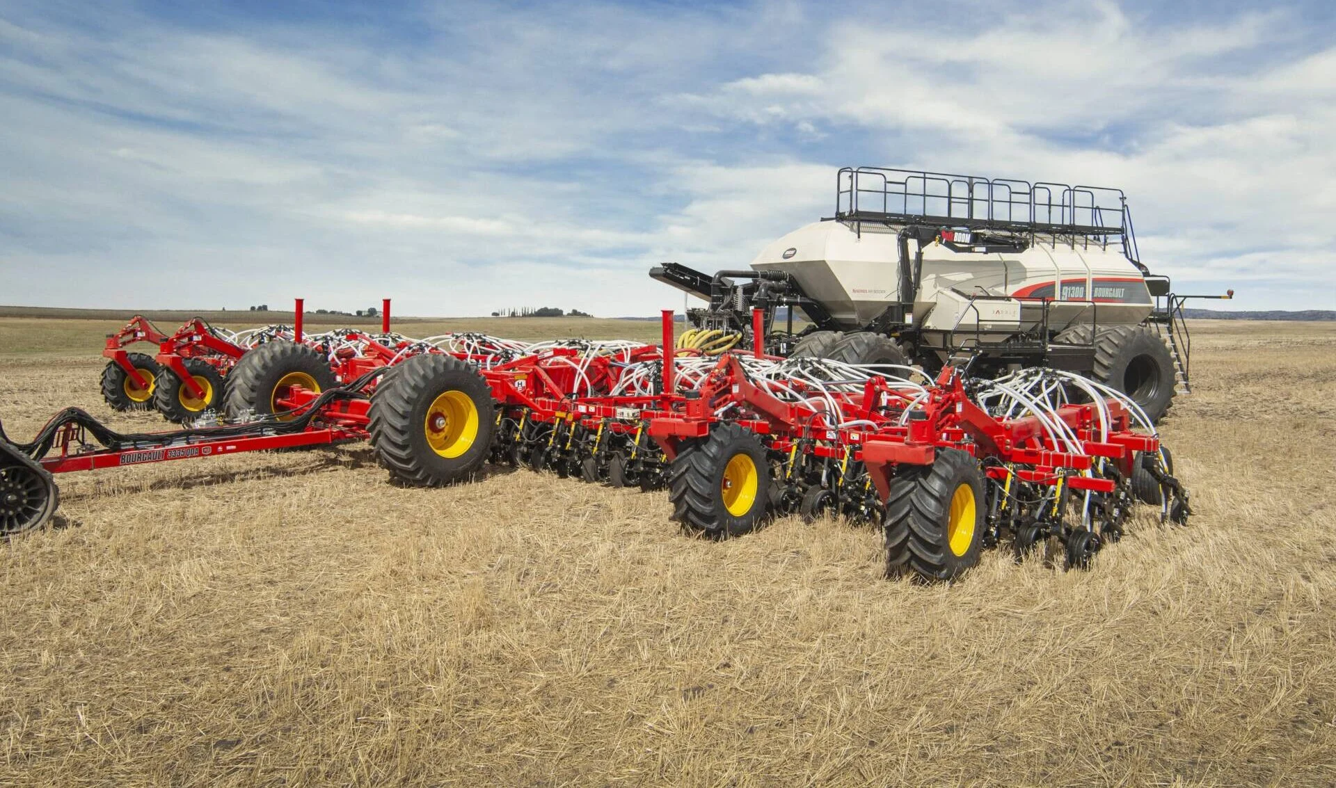 Large red agricultural planter attached to a white seed tank, positioned in a dry field under a partly cloudy sky.