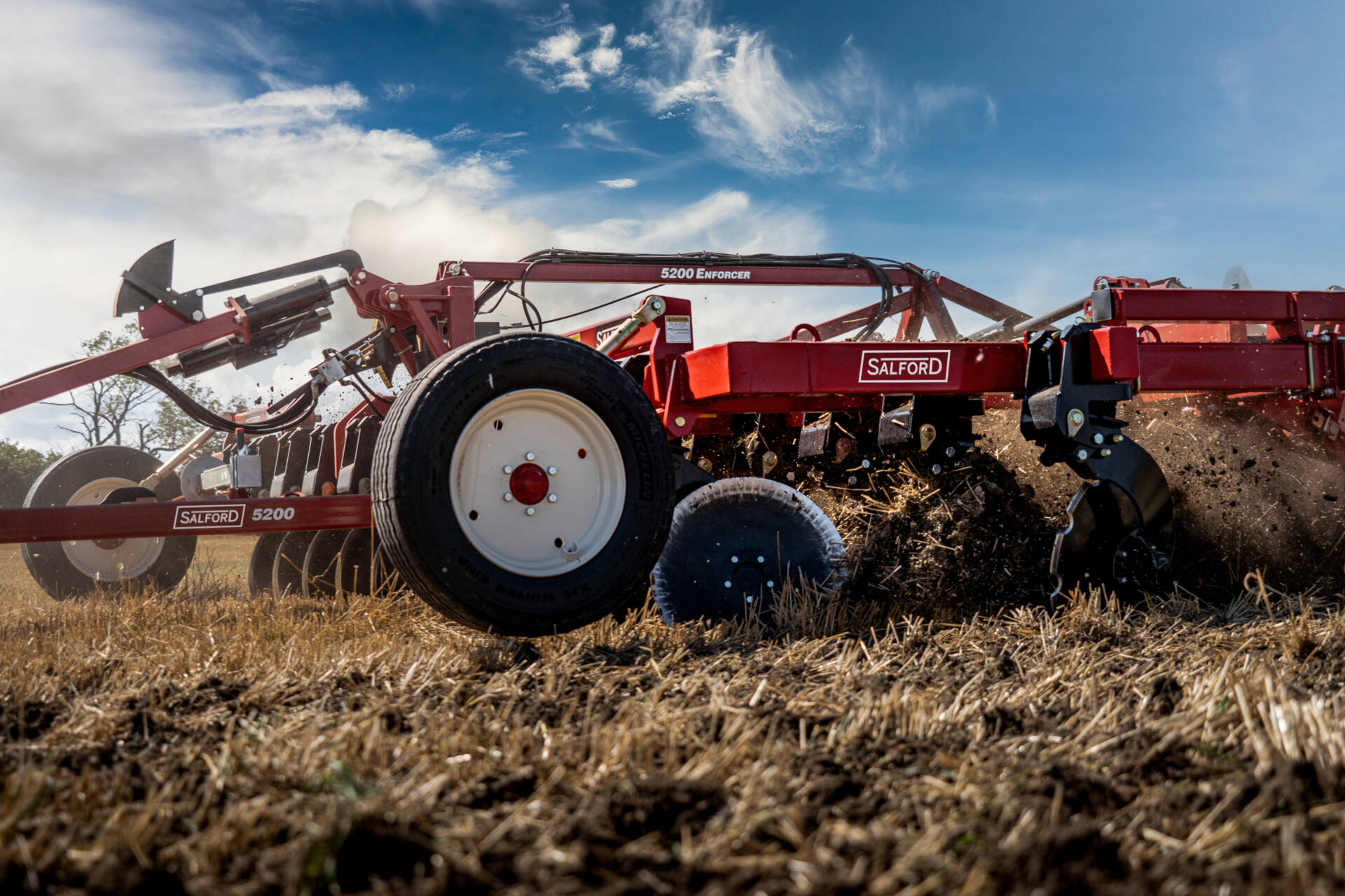 Close-up of a red agricultural cultivator tilling dry soil in a field under a partly cloudy sky.