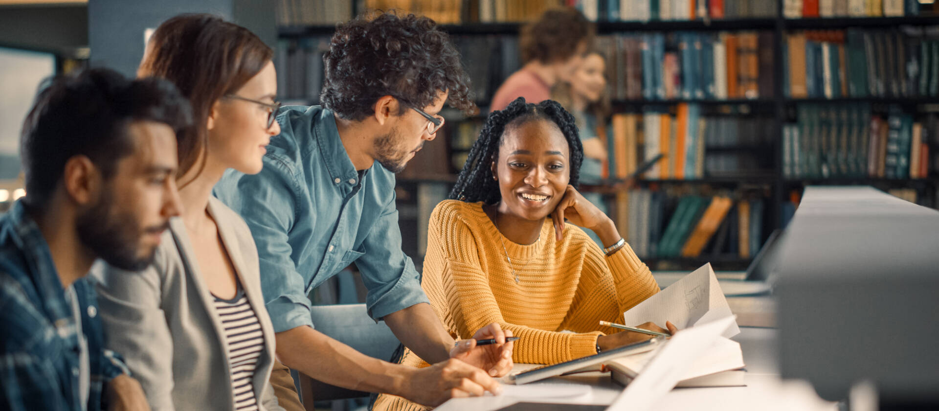 A diverse group of young adults collaborating and studying together in a modern library with laptops and books.