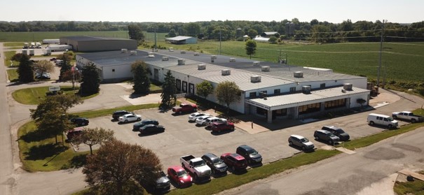 Aerial view of a commercial building with a parking lot filled with cars, surrounded by green fields and trees under a clear sky.