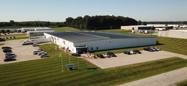 Aerial view of a large industrial warehouse surrounded by neatly trimmed grass and parked vehicles under a clear sky.