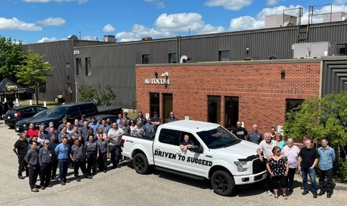 Group of people standing outside a brick building next to a white pickup truck with "Driven to Succeed" written on its side under a clear blue sky.