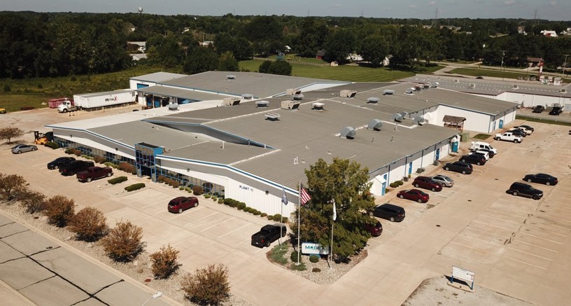 Aerial view of a large commercial building with multiple parked cars in the surrounding parking lots, situated near open green spaces.