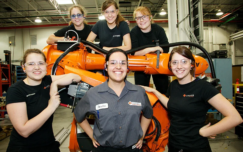 A group of six smiling women engineers posing with an industrial robotic arm inside a factory setting.