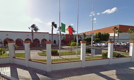 Exterior view of a government building with Mexican flags and a fenced courtyard under a clear sky.