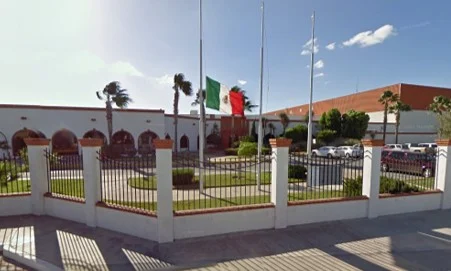 Exterior view of a government building with Mexican flags and a fenced courtyard under a clear sky.