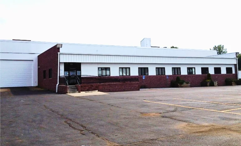 Large, single-story industrial building with white walls, red brick lower facade, multiple windows, and a gray roof beside an empty paved lot.