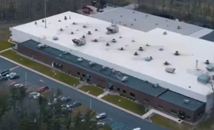 A large rectangular commercial building with a white roof and multiple rooftop units, surrounded by parked cars and greenery.
