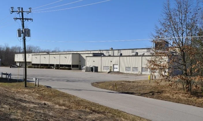 Large industrial warehouse with multiple loading docks, surrounded by pavement and sparse trees, under a clear blue sky.