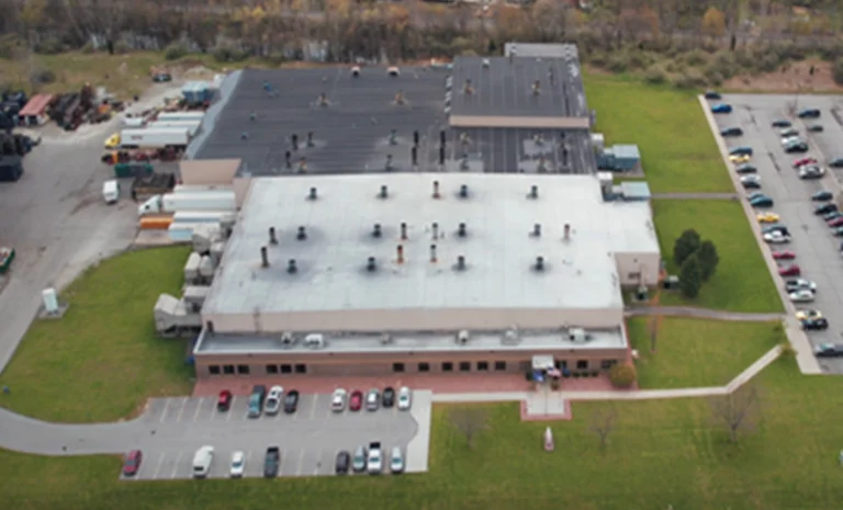 Aerial view of a large industrial building with multiple sections, surrounded by parked cars and green lawn.