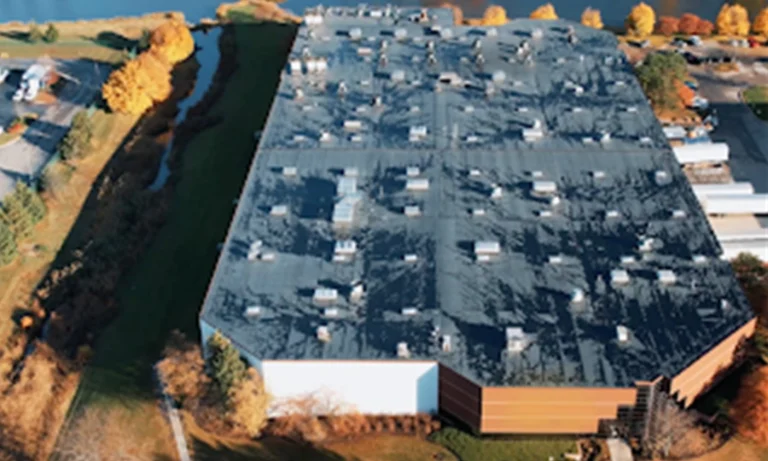 Aerial view of a large warehouse or industrial building with multiple rooftop units, surrounded by trees with autumn foliage.