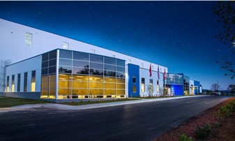 Modern industrial warehouse building with large glass windows illuminated at dusk against a clear evening sky.