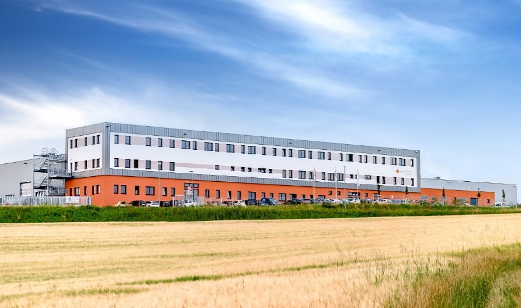 Modern industrial warehouse building with white and orange exterior situated near open fields under a blue sky.