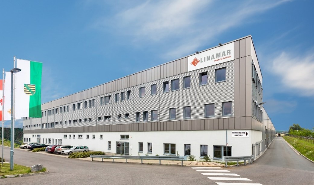 Modern industrial building with a white and gray exterior under a clear blue sky, featuring company signage and green landscaping.