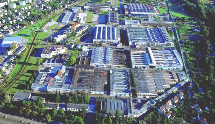 Aerial view of a large industrial complex featuring multiple factory buildings with solar panels on the roofs, surrounded by greenery and roads.