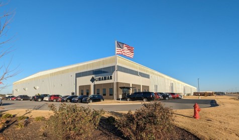 Exterior view of a modern industrial warehouse with an American flag flying above and multiple parked cars in front under a clear blue sky.