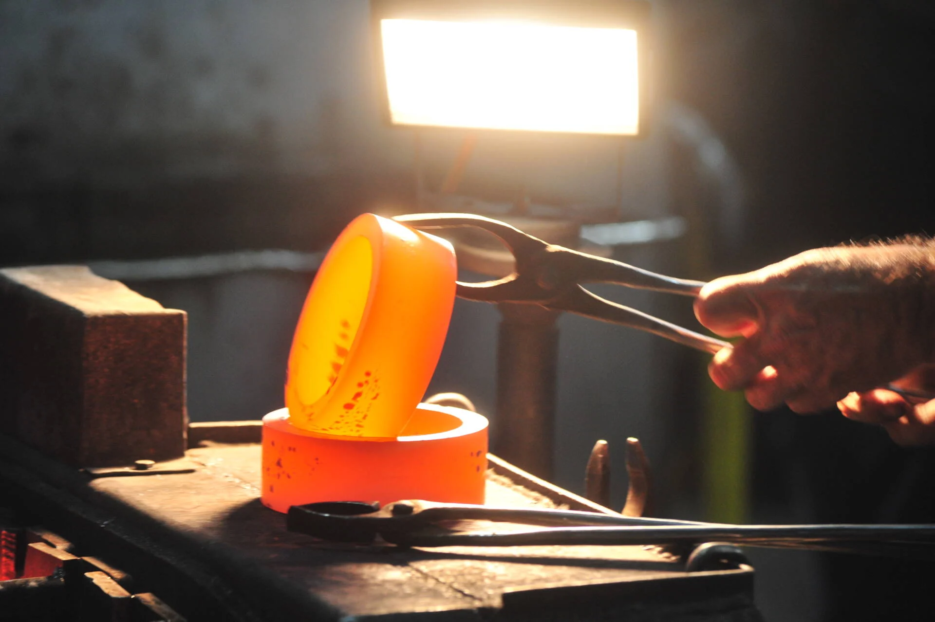 Close-up of a blacksmith using tongs to hold glowing hot metal shaped like a horseshoe on an anvil, illuminated by bright workshop lighting.