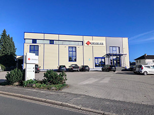 Exterior view of a modern beige commercial building with the Kubala logo, surrounded by parked cars and a clear blue sky.