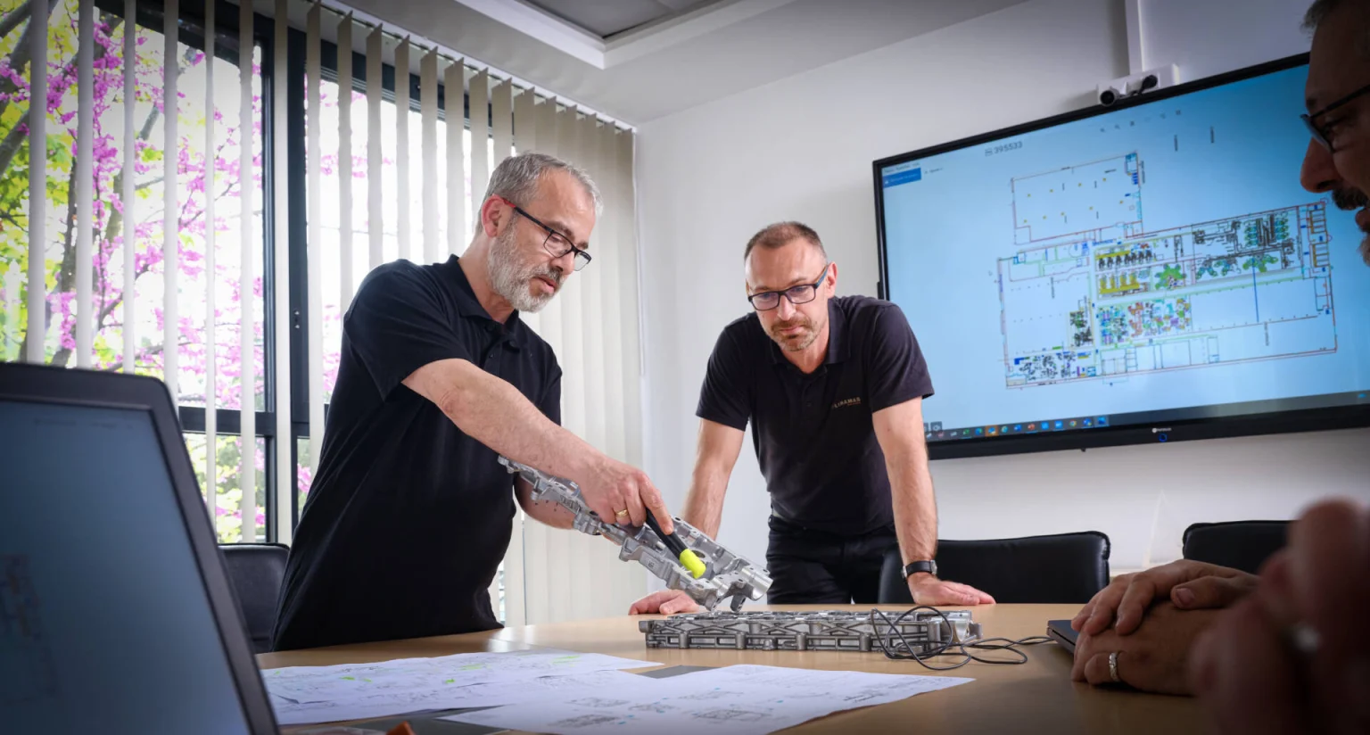 Two men collaborating on an architectural model in a modern office with blueprints and a digital floor plan displayed on a large screen.