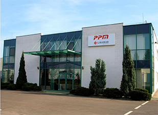 Modern commercial building with large glass windows and a sign reading "PPM" above the entrance, surrounded by neatly trimmed shrubs.