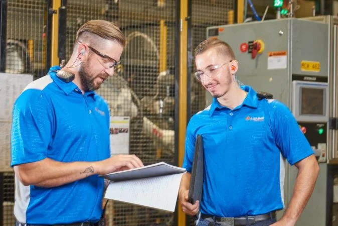 Two factory workers in blue shirts discussing notes on a clipboard inside an industrial facility.