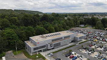 Aerial view of a modern commercial building surrounded by a large parking lot filled with cars and dense green trees in the background.