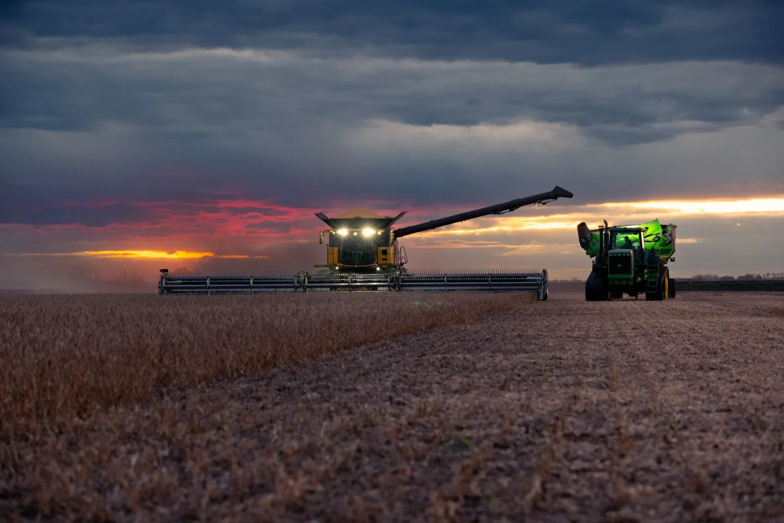 Combine harvester and tractor working together to harvest crops in a field during sunset under a cloudy sky.