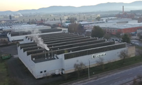 Aerial view of an industrial factory complex with multiple smokestacks emitting smoke, set against a mountainous background at sunset.