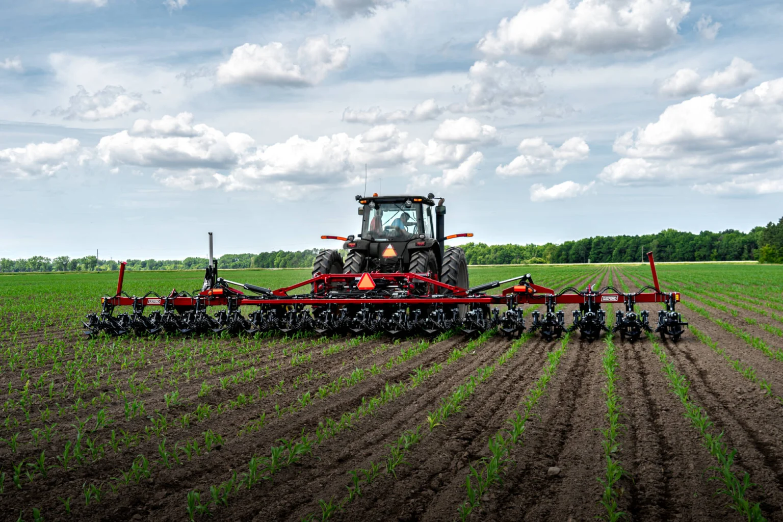 Tractor equipped with a wide cultivator tilling soil in a large agricultural field under a partly cloudy sky.