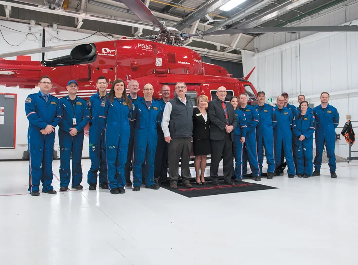 Group of pilots and crew members standing in front of a red rescue helicopter inside an aircraft hangar.