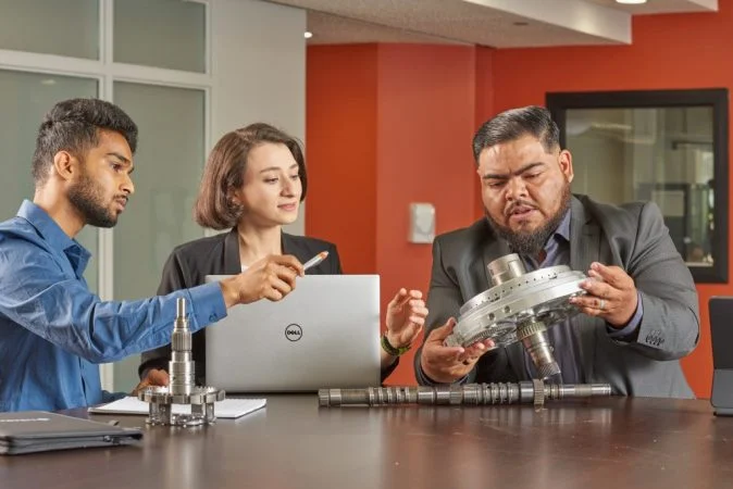 Three professionals in a meeting room examining a mechanical part while discussing technical details.